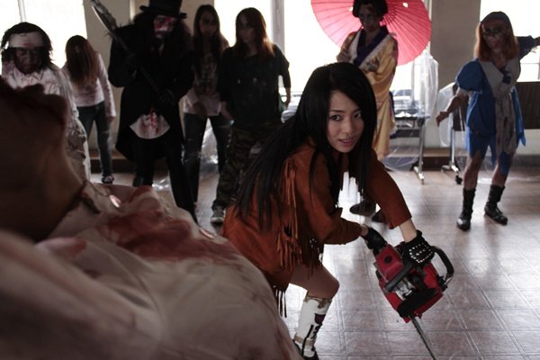 Lena, nuestra protagonista, armada con una motosierra. Al fondo, el peculiar grupo de zombies del que destaca la geisha con su sombrilla.